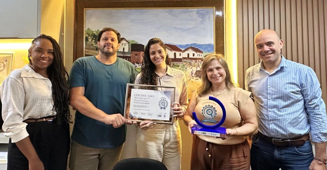 Close de cinco pessoas (três mulheres e dois homens) segurando o certificado de 100 pontos e o troféu Selo Diamante. Ao fundo, uma pintura em tela retratando casas rurais e uma parede com painel de madeira ripada. O clima é de celebração.