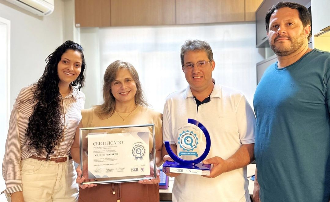 Registro em plano médio de quatro representantes da equipe da Prefeitura de Dores do Rio Preto. À esquerda, uma mulher de cabelos longos e cacheados e uma mulher loira seguram juntas o certificado emoldurado. Ao centro, um homem de camisa polo branca segura o troféu azul do 