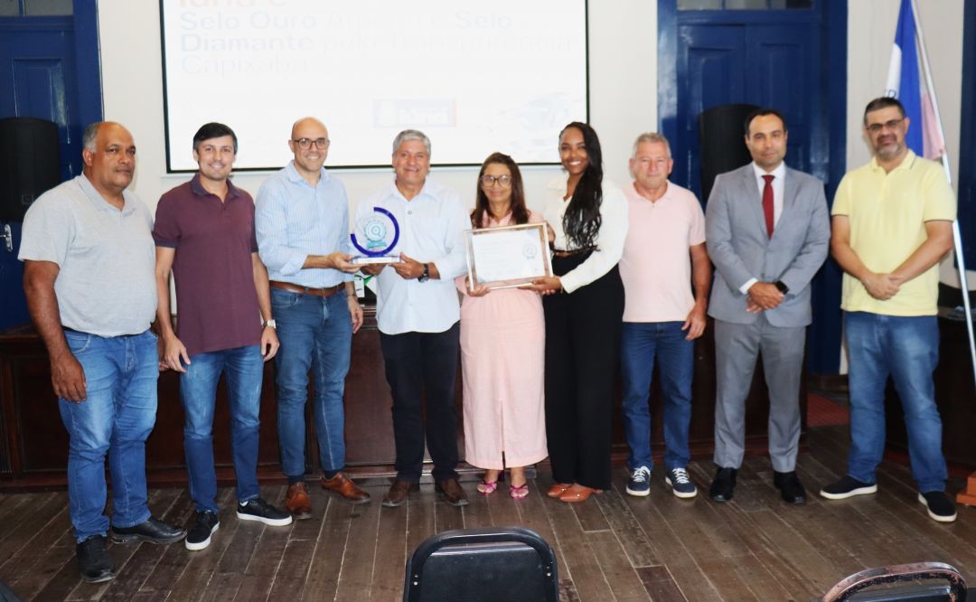 Fotografia em plano aberto mostrando um grupo de nove pessoas (sete homens e duas mulheres) no palco da Casa da Cultura de Iúna. Ao centro, o prefeito Romário Vieira e a secretária Elzeni Silva seguram, juntos com representantes do ES em Ação, o troféu azul e o certificado emoldurado do Selo Diamante.