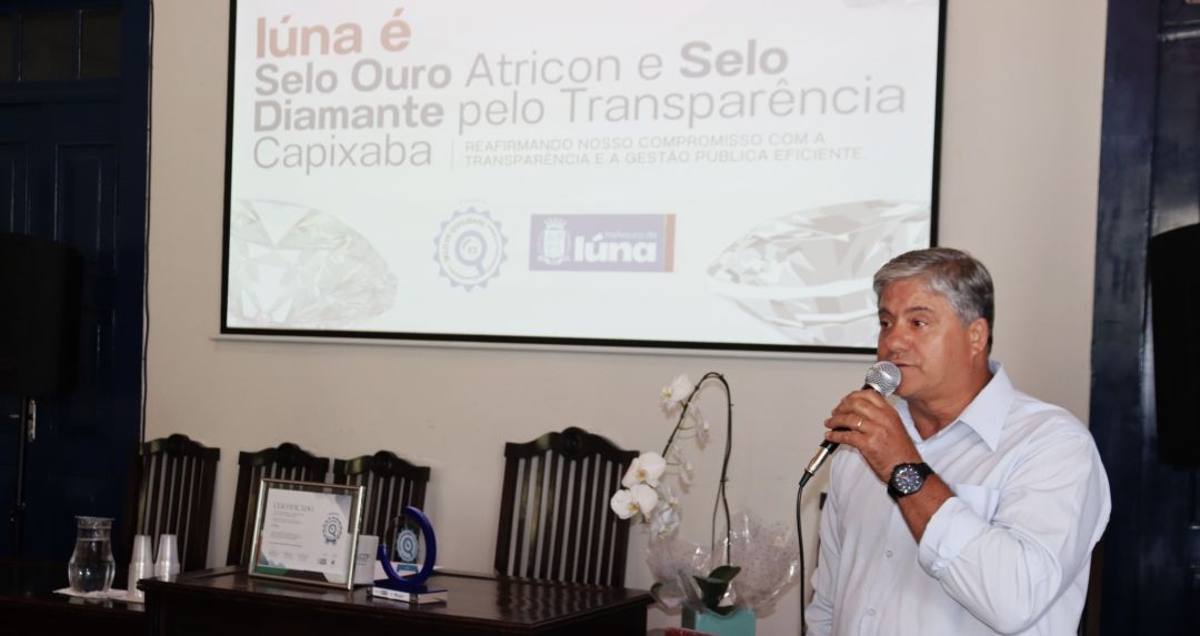 Prefeito Romário discursando durante solenidade de entrega do Selo Diamante de Transparência, tendo ao fundo um telão com a logo da Prefeitura e dizeres sobre a cerimônia.