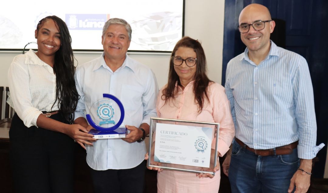 Foto aproximada de quatro pessoas segurando as premiações. Da esquerda para a direita: a coordenadora do ES em Ação, Débora Macedo, o prefeito Romário Vieira segurando o troféu circular azul, a secretária Elzeni Silva segurando o certificado emoldurado e o superintendente Luciano Golner. Ao fundo, o logo da Prefeitura de Iúna.