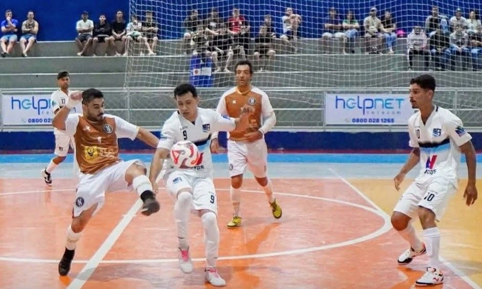 Momento de disputa intensa na segunda rodada da Copa Help. O jogador camisa 9, de branco, tenta proteger a bola contra a marcação de um atleta de laranja (Estrela Real x Top Motos). A imagem captura o dinamismo do futsal no Ginásio Romeu Rios.