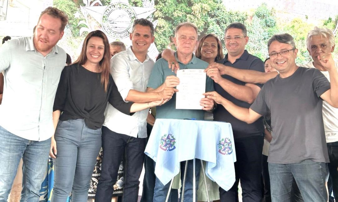 Fotografia colorida em plano médio mostrando o governador Renato Casagrande ao centro, segurando um documento oficial assinado ao lado de autoridades e lideranças políticas. Ao fundo, um painel com o brasão do Estado do Espírito Santo e a frase 