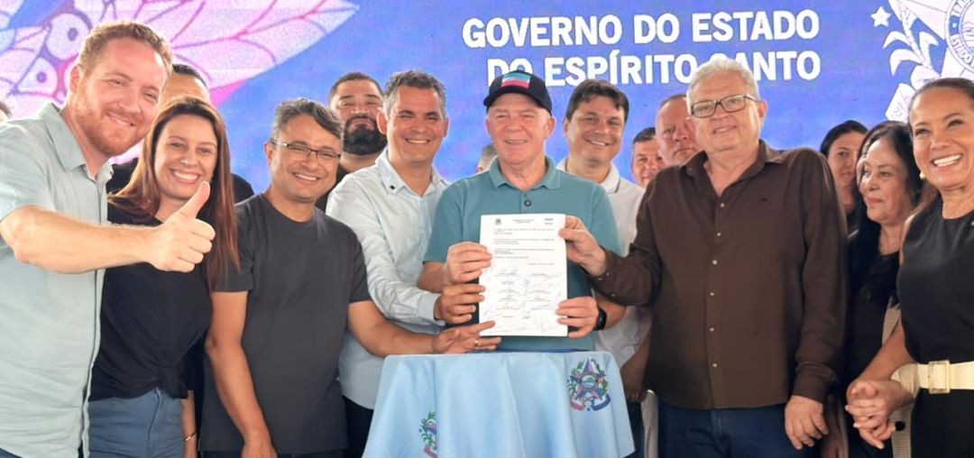 Fotografia colorida de mostrando um grande grupo de autoridades, incluindo o governador Renato Casagrande e o prefeito de Apiacá, segurando um documento assinado sobre uma mesa forrada com uma toalha azul com o brasão estadual. Ao fundo, um painel exibe uma foto com os dizeres 