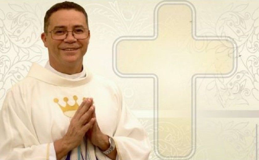 Fotografia do Padre José Carlos Ferreira da Silva sorrindo, usando óculos e vestes litúrgicas brancas (alva) com detalhes dourados. Ele está com as mãos postas em oração. Ao fundo, uma arte suave em tons de bege com o desenho de uma cruz estilizada e arabescos.