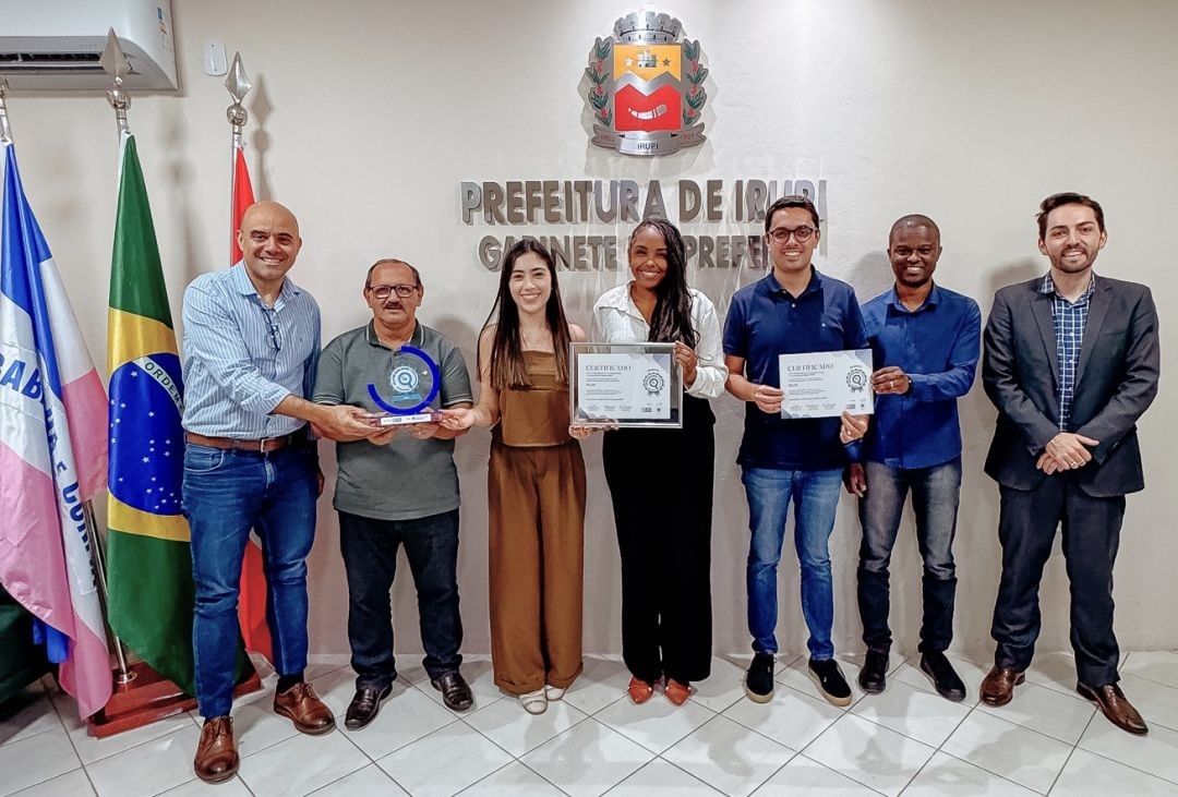 Fotografia no gabinete da Prefeitura de Irupi. Sete pessoas posam para a foto segurando o troféu circular azul do Selo Diamante e certificados de transparência pública. Ao fundo, o brasão do município e as bandeiras oficializadas.