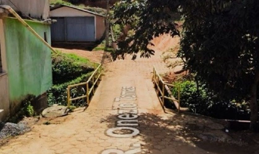 Fotografia de uma ponte estreita de calçamento em Irupi, com guarda-corpo amarelo, ligando ruas residenciais. No chão, lê-se parte do nome da Rua Onésio Tomaz.