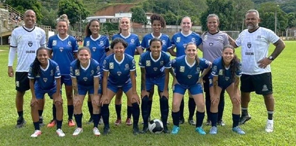 Fotografia colorida da equipe de futebol feminino de Colatina posando em um campo de grama verde. Elas vestem uniformes azuis com detalhes brancos. Ao lado das jogadoras, dois integrantes da comissão técnica vestem camisas brancas com a escrita 