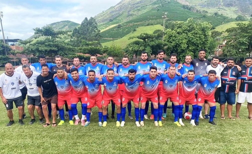 Fotografia do time de futebol masculino de Iúna posando em um campo gramado. Os jogadores vestem camisas com degradê do azul para o vermelho e calções vermelhos. Integrantes da comissão técnica usam camisas brancas com o nome 