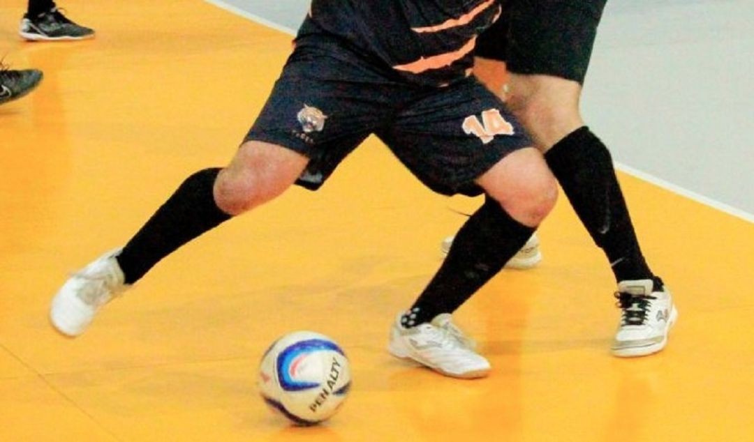 Fotografia em close das pernas de dois jogadores de futsal em disputa de bola. Um jogador veste uniforme preto com detalhes laranjas e número 14, enquanto o outro veste uniforme escuro. A bola, da marca Penalty, está no centro do piso esportivo amarelo do ginásio.