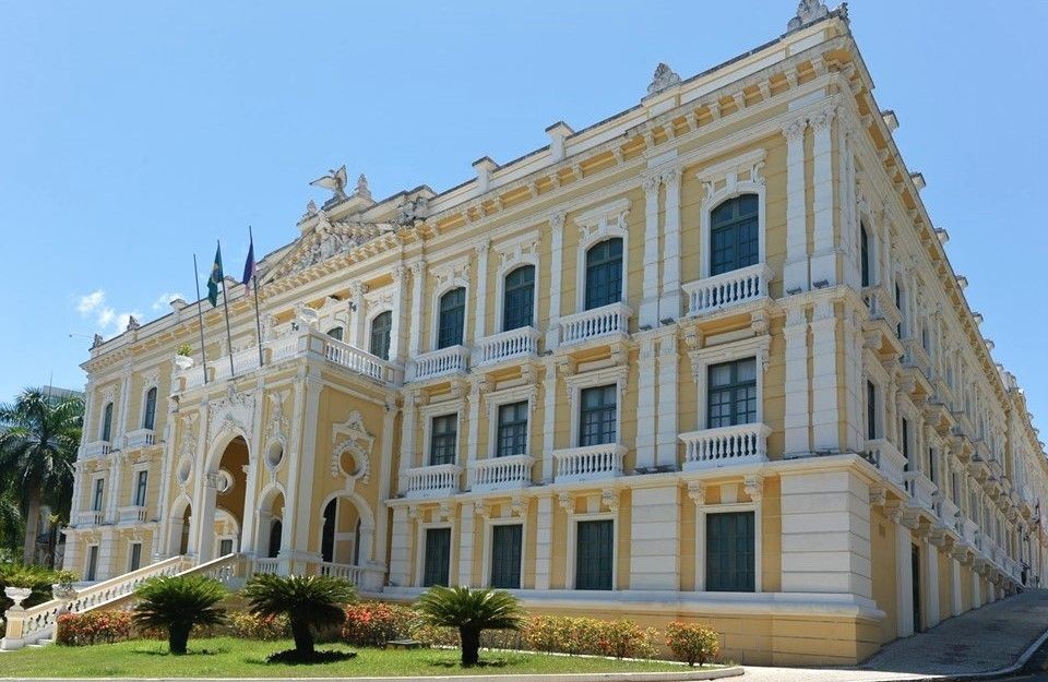 Fotografia da fachada lateral do Palácio Anchieta, sede do Governo do Espírito Santo, em Vitória. O prédio histórico possui arquitetura neoclássica em tons de amarelo e branco, com janelas emolduradas e detalhes em relevo. À frente, um jardim bem cuidado com palmeiras baixas e gramado verde sob um céu azul limpo. As bandeiras do Brasil e do Espírito Santo estão hasteadas na entrada.