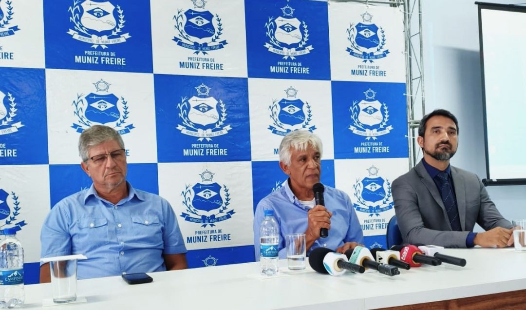 A imagem mostra o auditório do Cras em Muniz Freire durante uma coletiva de imprensa. Três homens estão sentados à mesa: à esquerda, o secretário Marciano Areias, de camisa azul e óculos; ao centro, o prefeito Dito Silva, de cabelos brancos, falando ao microfone; e à direita, o procurador Marcos Valério, vestindo paletó cinza e gravata azul. À frente deles, vários microfones de imprensa captam o áudio. O fundo é composto por um painel xadrez em azul e branco com o brasão oficial da Prefeitura de Muniz Freire.