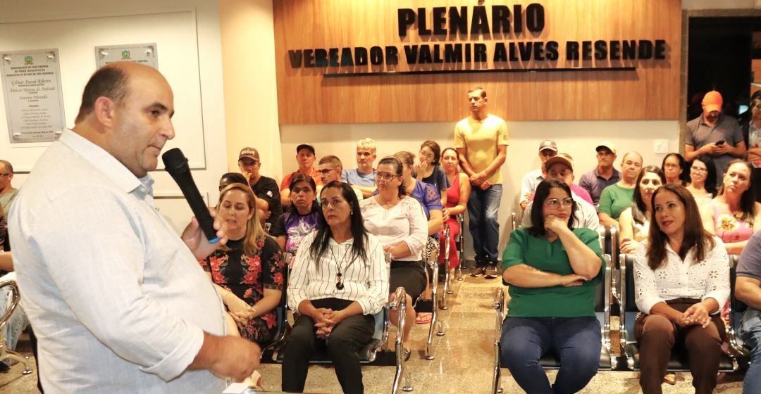 Prefeito Dudu Queiroz fala ao microfone a pessoas presentes à reunião social sobre o início das obras da ES-493 que estão sentadas em cadeiras, tendo ao fundo o painel que identifica o 