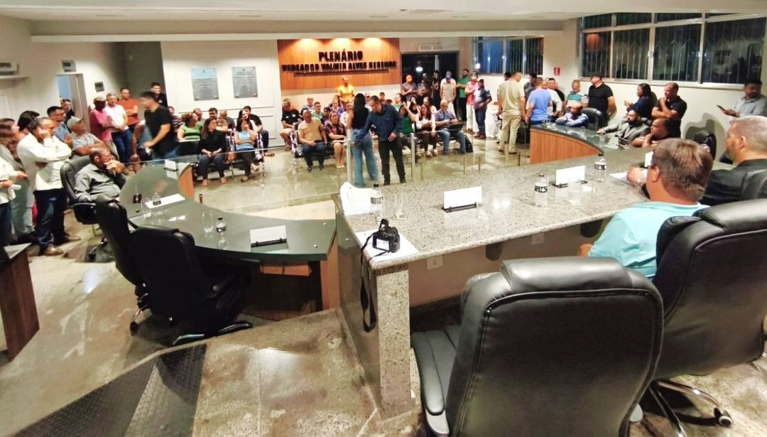 Visão panorâmica do plenário da Câmara Municipal de Divino de São Lourenço, lotado de cidadãos e autoridades acompanhando a apresentação do projeto rodoviário. Ao fundo, painel com o nome do plenário e uma projeção técnica na parede.