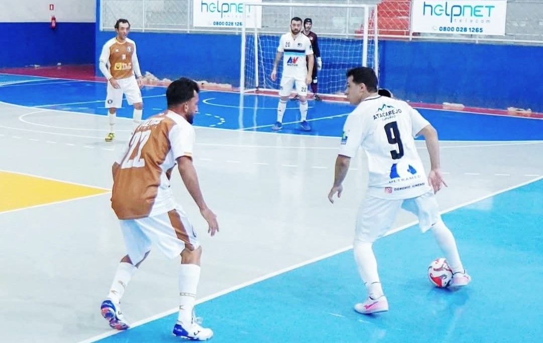 Fotografia de uma partida de futsal em quadra azul e cinza. Em primeiro plano, dois jogadores disputam a posse de bola: um de uniforme branco com o número 9 nas costas e outro de uniforme branco e cobre com o número 22. Ao fundo, o goleiro e outros jogadores observam a jogada perto da trave. Placas de publicidade da 