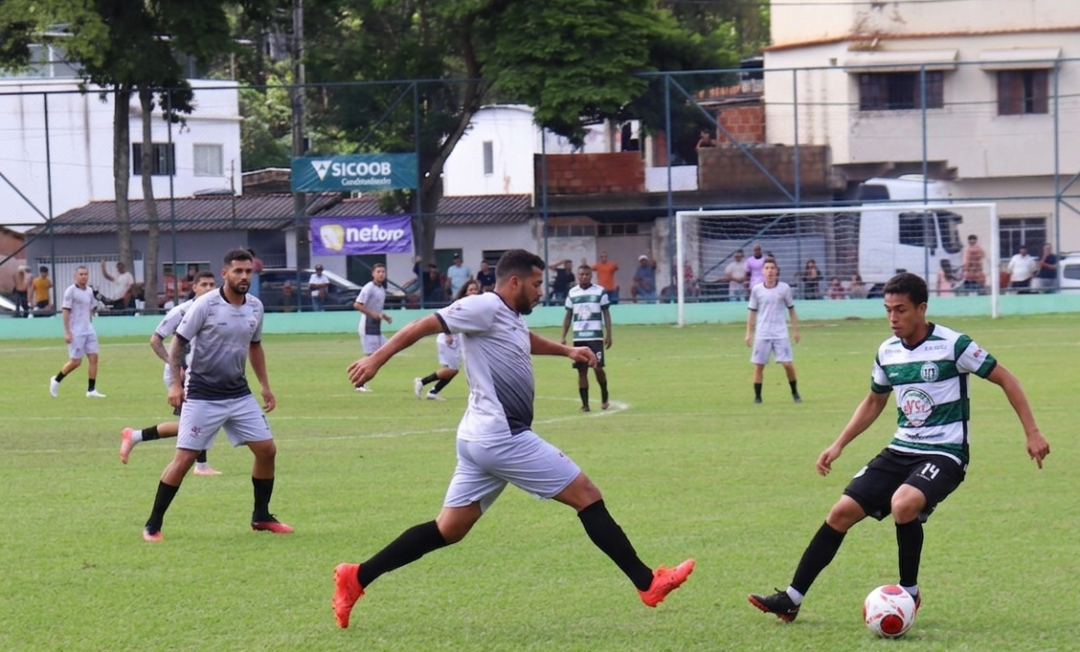 Fotografia de uma partida de futebol de campo em um dia ensolarado. No centro, jogadores disputam a posse de bola no gramado verde. À direita, um jogador com a camisa listrada em verde e branco disputa bola com outro de cinza e preto. Outros jogadores que vestem uniforme cinza e preto e de verde e branco observam. Ao fundo, vê-se a rede do gol, alambrados com faixas de patrocinadores como Sicoob e NetDRP, árvores altas e construções urbanas simples.