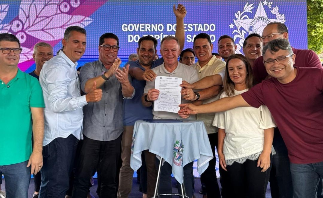 Foto colorida mostra o governador Renato Casagrande ao centro, cercado por lideranças políticas e secretários estaduais. Ele segura e exibe um documento assinado (ordem de serviço) sobre uma mesa redonda forrada com uma toalha azul clara que leva o brasão do Espírito Santo. Ao fundo, um painel de LED exibe a inscrição 