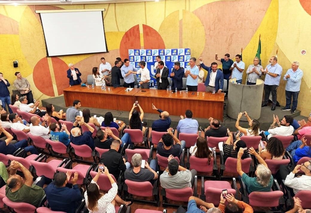 Imagem em plano geral de cima para baixo, mostrando um grande público sentado em poltronas vermelhas numa plateia. Ao fundo, num palco com mesa comprida de madeira, várias figuras políticas de pé posam para o anúncio. Há uma tela de projeção branca no alto e um painel azul e branco com o logo 