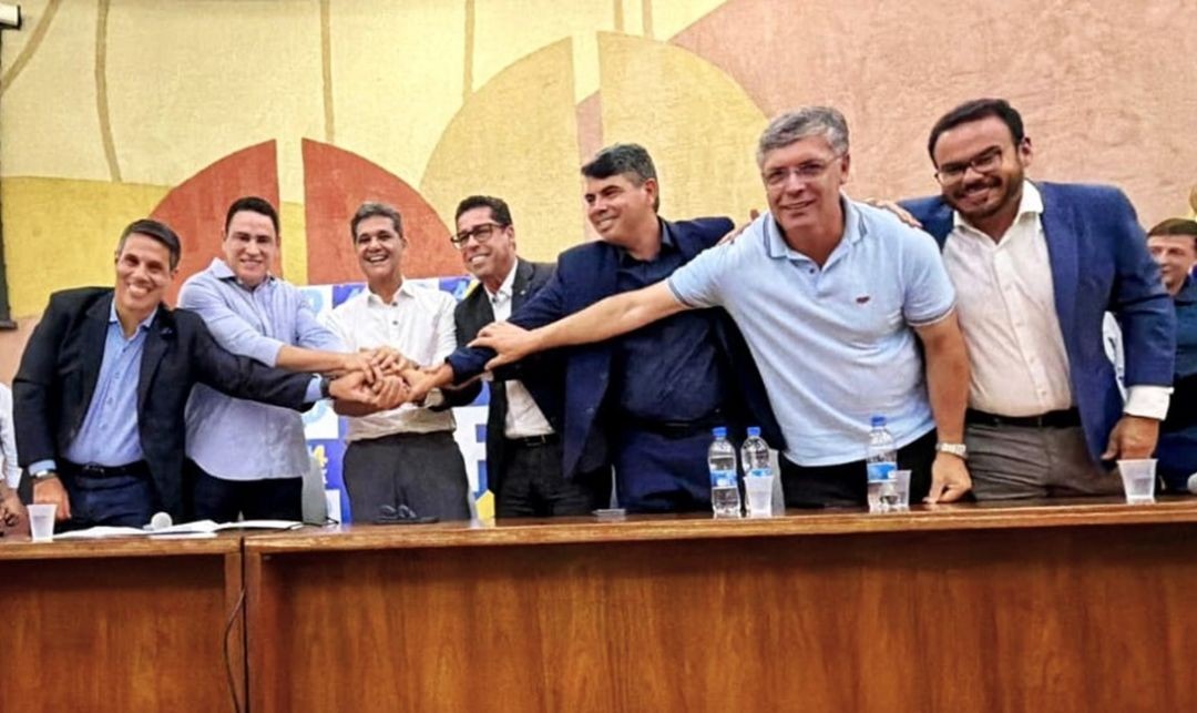 Imagem em plano médio horizontal, capturando um momento de cooperação e unidade no palco. No centro, seis homens, incluindo Ricardo Ferraço e os deputados Da Vitória e Marcelo Santos, posam de pé atrás de uma mesa, com as mãos sobrepostas num gesto simbólico de apoio e aliança. Todos sorriem para a câmera, vestidos com ternos e camisas sociais. Ao fundo, vê-se parte do painel do União Brasil e garrafas de água sobre a mesa.
