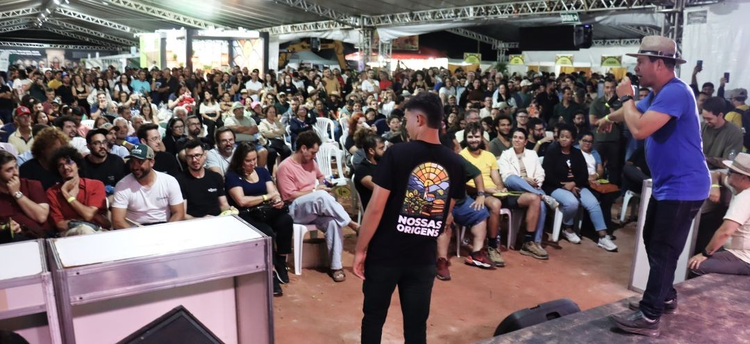 Fotografia de um evento em local coberto por tenda branca. No palco, à direita, um homem de camiseta azul e chapéu fala ao microfone. Diante dele, uma plateia sentada em cadeiras brancas acompanha a apresentação. No centro, um homem de costas veste uma camiseta preta com a estampa 
