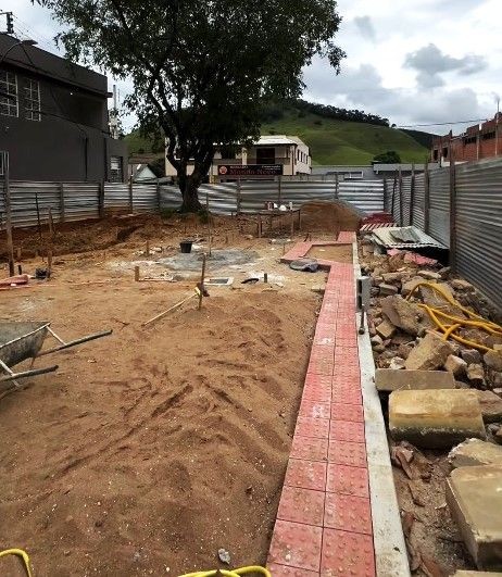 Área em construção cercada por tapumes metálicos. O chão é de terra com areia, e um novo caminho de piso tátil vermelho já começou a ser instalado. Ao centro, uma árvore de grande porte preservada e, ao fundo, montanhas e o comércio local.
