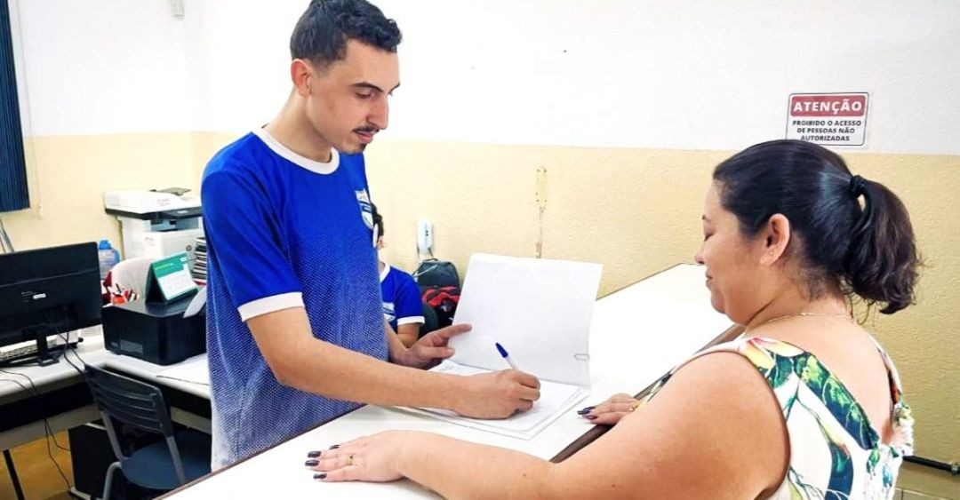 Em um ambiente de escritório, um jovem de camisa azul assina documentos sobre um balcão branco. À direita, uma mulher de blusa florida acompanha o procedimento. Ao fundo, há uma placa de aviso com a inscrição 