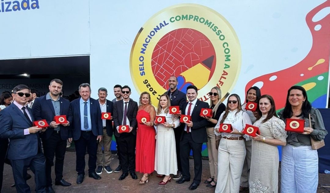 Representantes municipais e autoridades educacionais posam para foto oficial em Brasília. Eles seguram estojos vermelhos contendo o Selo Ouro de Alfabetização. Ao fundo, um painel circular amarelo e vermelho com o logotipo do programa 