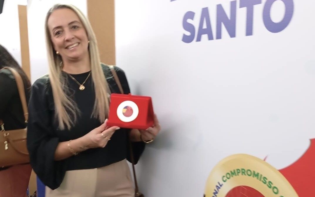 A secretária de Educação de Ibitirama, Laudiceia Zamboti de Souza, sorri enquanto segura o estojo vermelho com a medalha do Selo Ouro. Ao fundo, detalhe de um painel branco.