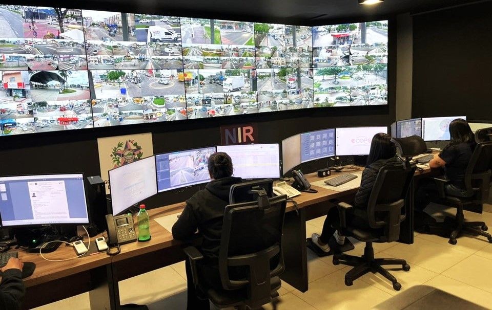Fotografia de uma central de monitoramento moderna. Em primeiro plano, três operadores trabalham sentados em cadeiras de escritório pretas diante de bancadas de madeira com vários monitores. Ao fundo, uma parede inteira é composta por um telão gigante dividido em dezenas de janelas exibindo imagens de câmeras de segurança de vias públicas, cruzamentos e calçadões em tempo real. No centro da parede, abaixo do telão, lê-se a sigla 