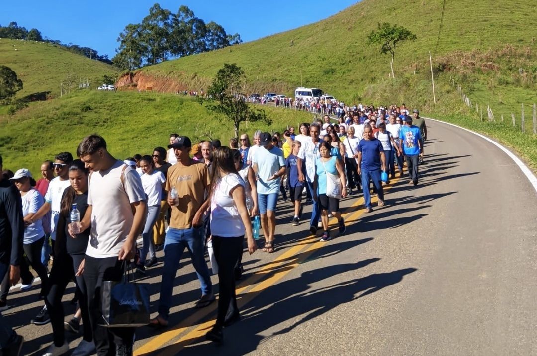 Fotografia em plano aberto mostra centenas de pessoas caminhando pelo acostamento de uma estrada asfaltada em Dores do Rio Preto. O grupo é composto por homens, mulheres e jovens. Ao fundo, veem-se montanhas verdes, pastagens e um ônibus acompanhando a procissão sob um céu azul.