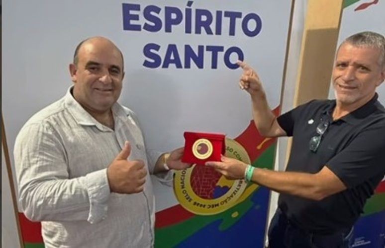 O prefeito Dudu (à esquerda) faz sinal de positivo e o secretário de educação, Luiz Carlos (à direita), aponta para o Selo Ouro do MEC, uma medalha dourada em estojo de veludo vermelho. Ao fundo, banner com a inscrição 