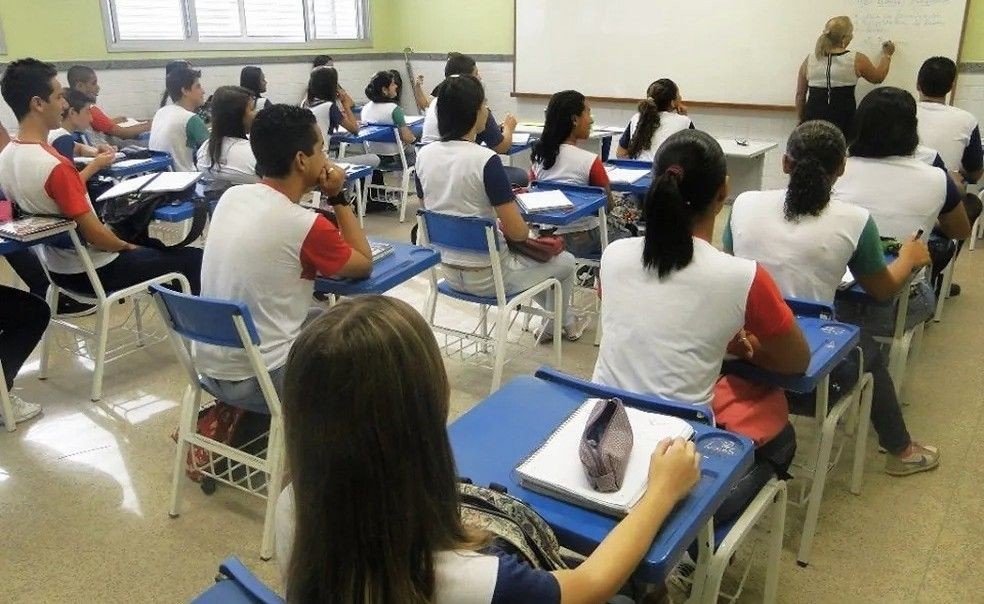 Fotografia de uma sala de aula com alunos adolescentes sentados em carteiras azuis, vestindo uniformes brancos e azuis ou brancos e vermelhos. Ao fundo, uma professora escreve em um quadro branco.