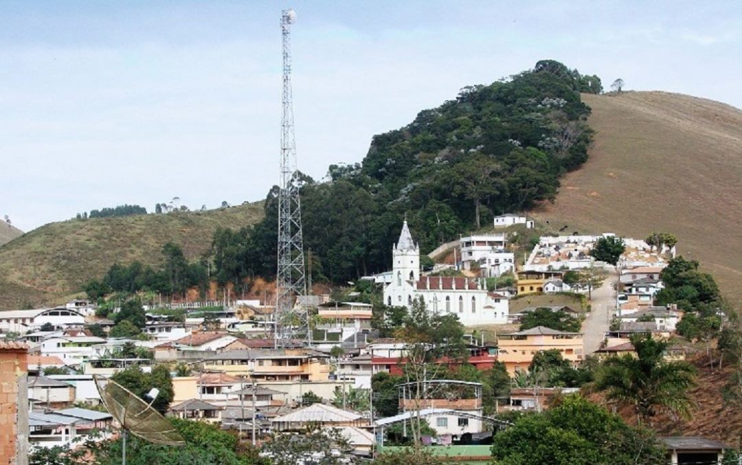 Foto de imagem geral da cidade de Divino de São Lourenço, tendo ao centro a torre de telefonia, no meio de casas e prédios, incluindo a Igreja Católica, tendo ao fundo uma mata e morros.