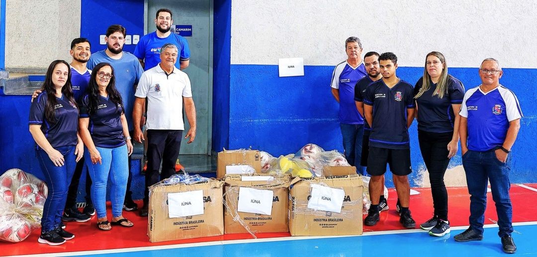Equipe da Secretaria de Esportes de Iúna, junto com o prefeito Romário Vieira, posando ao lado do material esportivo recebido que estão em caixas de papelão no centro da imagem, além de bolas de futebol, no canto esquerdo da foto.