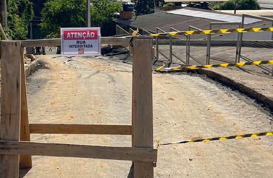 A imagem mostra um bloqueio de rua em uma área de aclive, utilizando um cavalete de madeira e fitas de sinalização zebradas (preta e amarela). Uma placa de aviso está fixada no cavalete, no canto superior esquerdo,
com as seguintes inscrições: 