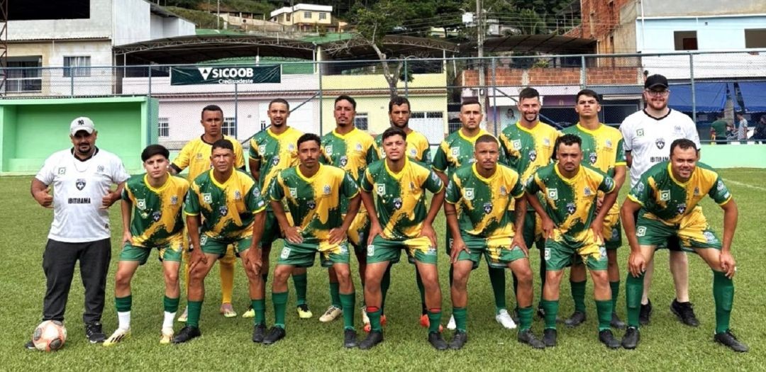 Fotografia oficial da equipe masculina de Ibitirama. Os jogadores posam uniformizados com camisas em tons de verde e amarelo, com detalhes gráficos em zigue-zague. Estão perfilados em duas fileiras (em pé e agachados) em um campo de grama natural. Ao fundo, árvores e construções da cidade.