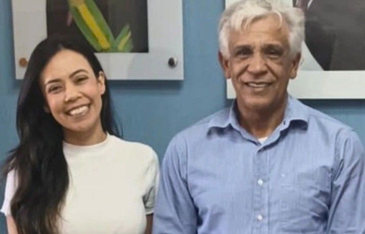 A diretora executiva do Consórcio Caparaó, Bruna Loura, e o prefeito de Muniz Freire, Dito Silva, posam lado a lado, sorrindo, em ambiente interno com parede azul e quadros ao fundo.