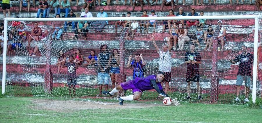 Goleiro se estica para defender cobrança de pênalti durante partida de futebol, com torcedores ao fundo em arquibancada.