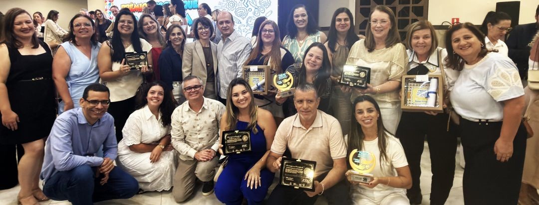Foto em grupo com cerca de 20 profissionais da educação em um salão de eventos. Algumas pessoas estão em pé e outras agachadas à frente. Vários integrantes seguram placas de homenagem e troféus dourados, celebrando a conquista do Selo Ouro em um clima de confraternização. Ao fundo, um painel colorido com elementos gráficos escolares.