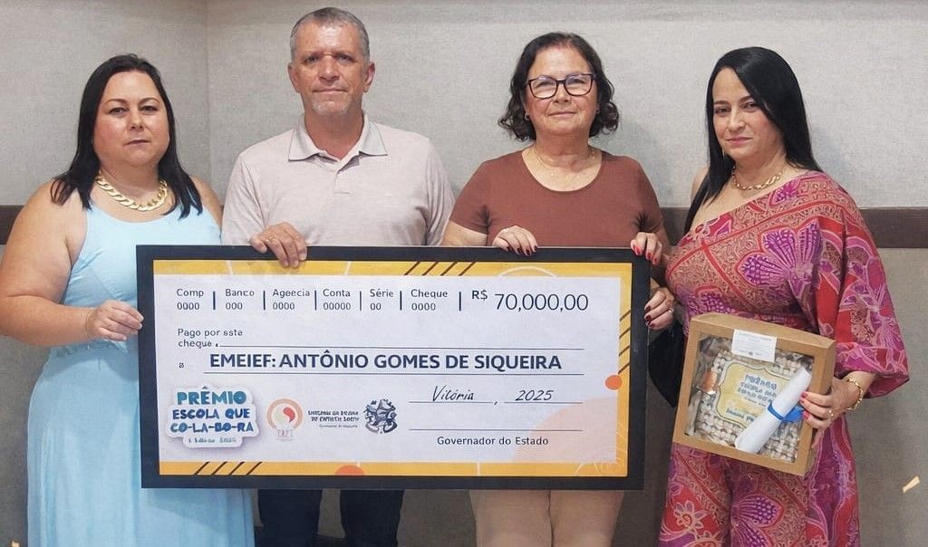 Fotografia em plano médio de quatro pessoas (duas mulheres nas extremidades, o secretário Luiz Carlos de Souza e uma mulher ao centro) segurando um cheque simbólico gigante. O cheque exibe o valor de R$ 70.000,00 e o nome da 