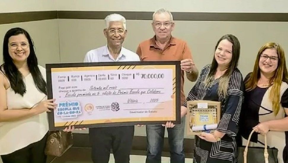 Cinco pessoas sorridentes seguram um cheque simbólico gigante no valor de 70 mil reais. À esquerda, duas mulheres; ao centro, dois homens (um de camisa branca e outro de camisa polo laranja); à direita, duas mulheres, uma delas segurando uma caixa de presente e um diploma. O cheque exibe a logomarca do Governo do Estado do Espírito Santo e do Prêmio Escola que Colabora.