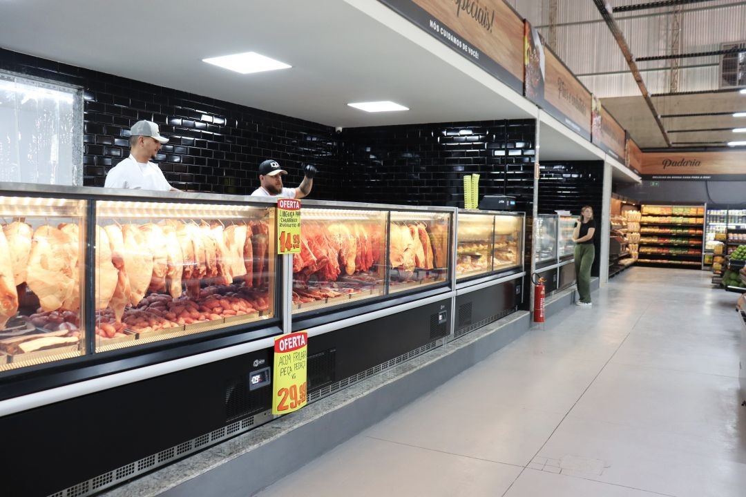 Uma visão detalhada do setor de açougue do Café Supermercados durante a inauguração. Dois açougueiros uniformizados e de boné estão atrás de um longo balcão refrigerado de vidro, que exibe diversos cortes de carne fresca, incluindo peças inteiras penduradas e embutidos. O balcão tem preços promocionais amarelos e pretos em destaque. Ao fundo, as paredes são revestidas com azulejos pretos brilhantes, e uma cliente observa os produtos. A iluminação embutida no teto é clara e moderna, e a identificação do setor é visível em um painel superior.