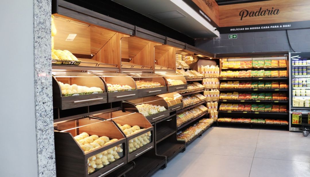 O moderno setor de padaria do Café Supermercados em Irupi. A imagem foca em expositores de madeira de alta qualidade com prateleiras de vidro e iluminação LED interna, repletos de pães frescos, pães de queijo e doces. Ao lado, gôndolas de aço exibem pães de forma embalados. Acima, um painel de madeira com a inscrição 