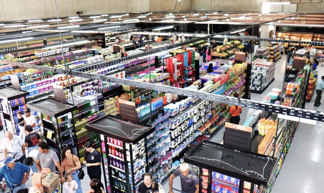 Visão aérea do interior do Café Supermercados. O local é amplo, bem iluminado e organizado com diversas gôndolas repletas de produtos. Há uma grande movimentação de clientes circulando pelos corredores com carrinhos de compra. O teto alto possui estrutura metálica e sistema de climatização aparente.