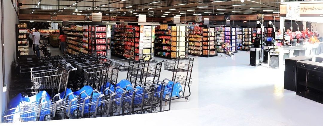 Visão do interior do Café Supermercados. O local é amplo, bem iluminado e organizado com diversas gôndolas repletas de produtos. O teto alto possui estrutura metálica e sistema de climatização aparente.