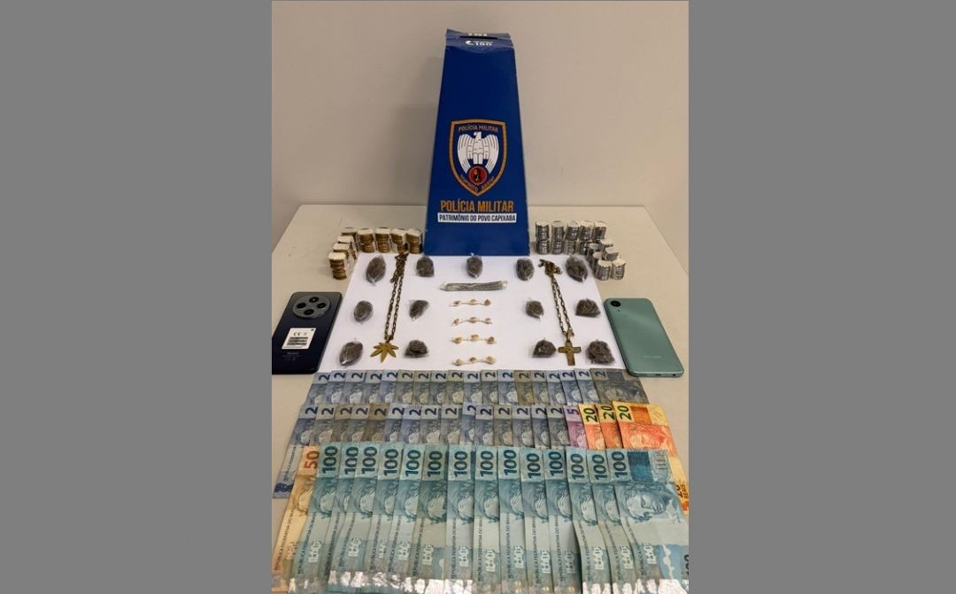 Mesa com materiais apreendidos pela Polícia Militar, incluindo dinheiro em notas, buchas de maconha, pedras de crack, celulares e itens utilizados para embalo de drogas, dispostos ao lado de um suporte com identificação da corporação.