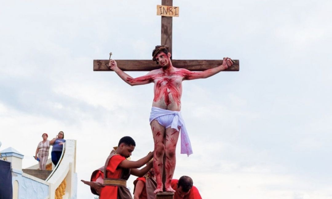 Homem representando Jesus Cristo crucificado durante encenação da Paixão de Cristo, com maquiagem simulando ferimentos, enquanto participantes caracterizados como soldados atuam ao redor em espaço aberto.