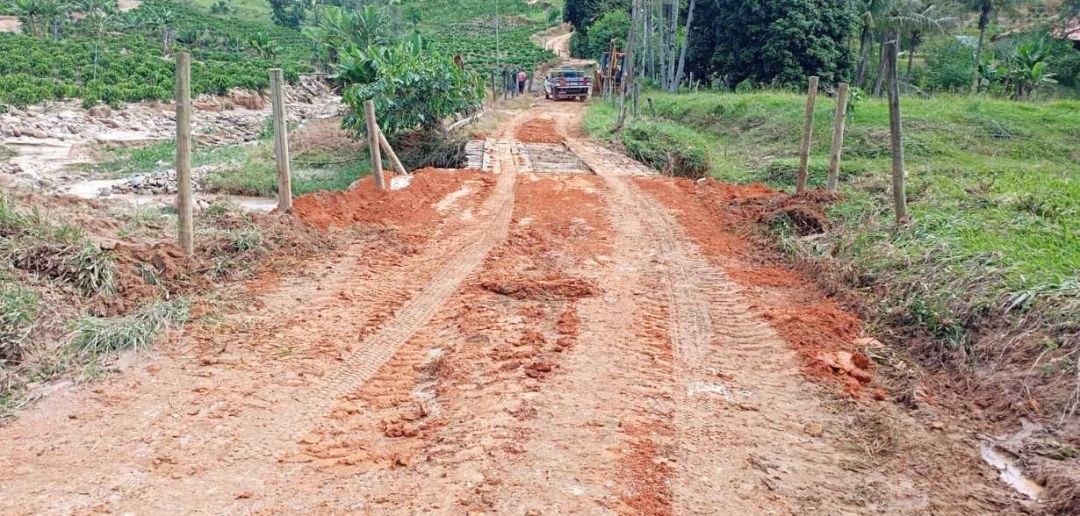 Estrada de terra em processo de recuperação, apresentando marcas de pneus de máquinas pesadas e aterro de solo avermelhado para nivelar o acesso à ponte.