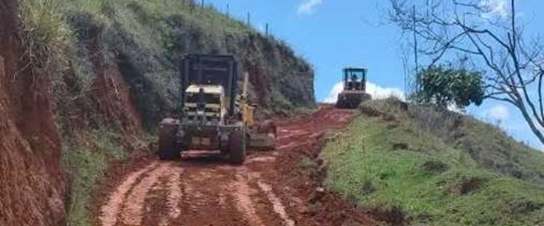 Máquinas pesadas da Prefeitura, incluindo uma motoniveladora (patrol), operam na manutenção de uma estrada de terra íngreme em Muniz Freire. O solo está úmido e apresenta marcas profundas de pneus, enquanto as máquinas realizam o nivelamento da via para restaurar a passagem após as chuvas.