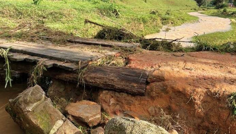 Detalhe da cabeceira de uma ponte de madeira danificada pela erosão. A força da água escavou a terra sob as vigas de sustentação, deixando a estrutura instável. Ao fundo, observa-se um curso d'água com volume elevado e cor de barro, cortando a vegetação rasteira.
